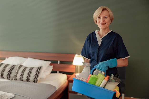 Empregada doméstica de uniforme para limpar apartamento segura caixa com detergentes e panos e olhando a câmera