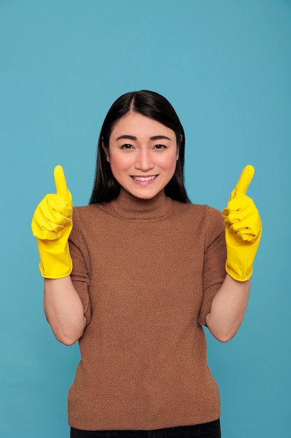 Empregada feliz e alegre animada do trabalho doméstico polegares para cima e usando luvas amarelas para segurança das mãos isoladas em um fundo azul, conceito de limpeza em casa, fêmea de clareira satisfeita com humor positivo