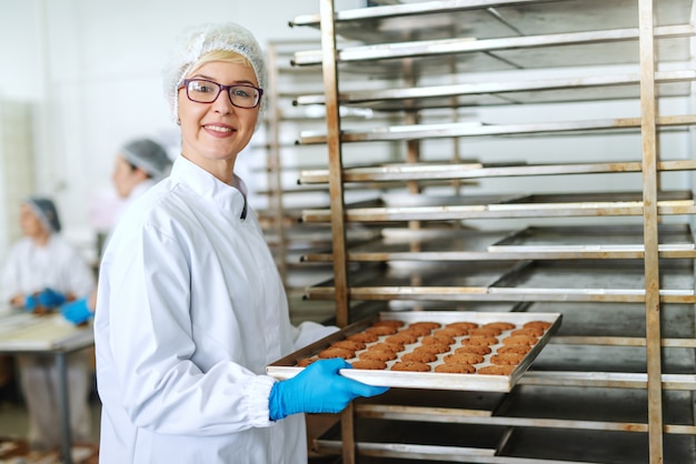 Empregada loira sorridente com óculos e em uniforme estéril, colocando a bandeja com deliciosos biscoitos na prateleira.