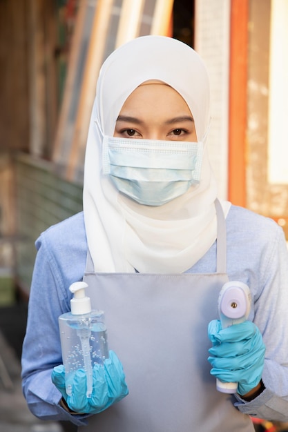 Empregada muçulmana ou lojista segurando álcool gel dispensador desinfetante para as mãos e pistola de scanner térmico para verificação de temperatura novo conceito de prática de negócios normal