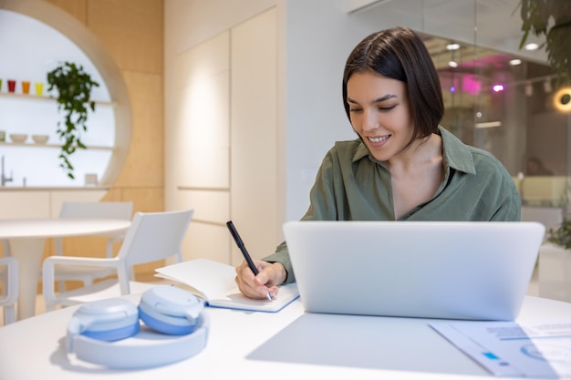 Empregada satisfeita da empresa sentada na tela do laptop escrevendo com uma caneta em seu diário