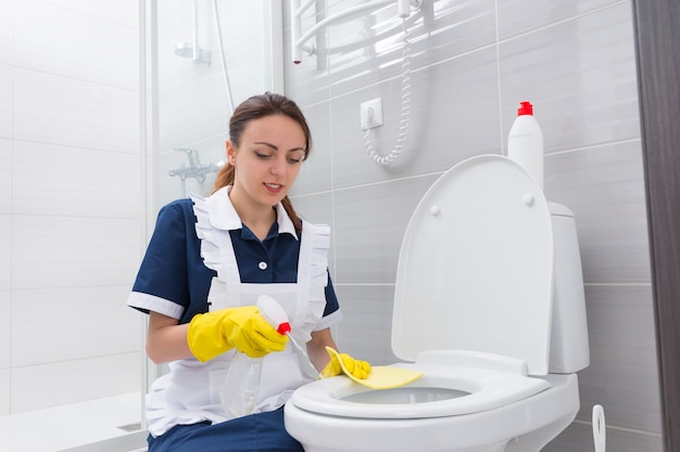 Empregada solteira com expressão alegre ajoelhada no vaso sanitário enquanto limpa o assento com spray e pano amarelo nas mãos enluvadas de borracha
