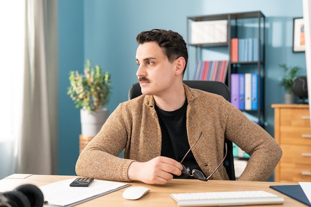 Empregado caucasiano de óculos sentado à mesa distraído do trabalho do computador ponderando jovens pensativos