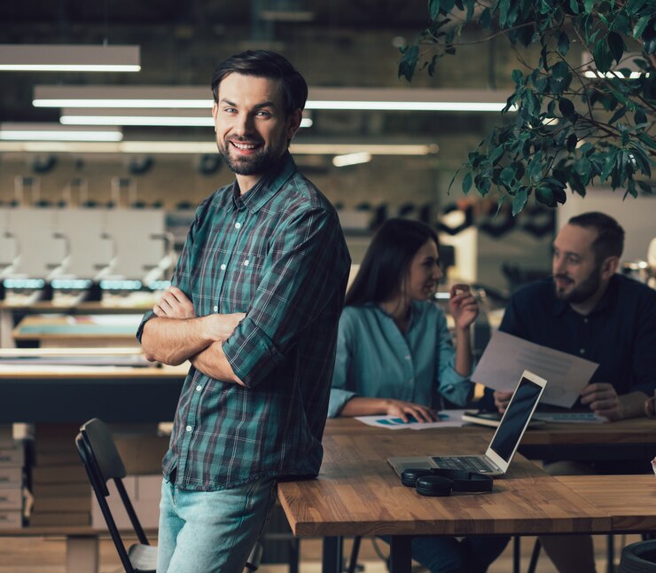 Empregado da editora alegre em pé com os braços cruzados e sorrindo. Homem e mulher calmos sentados na mesa conversando ao fundo