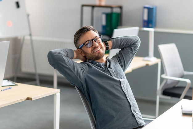 Empregado da empresa de meia idade confiante, apoiando-se na cadeira com as mãos atrás da cabeça, relaxando e sorrindo