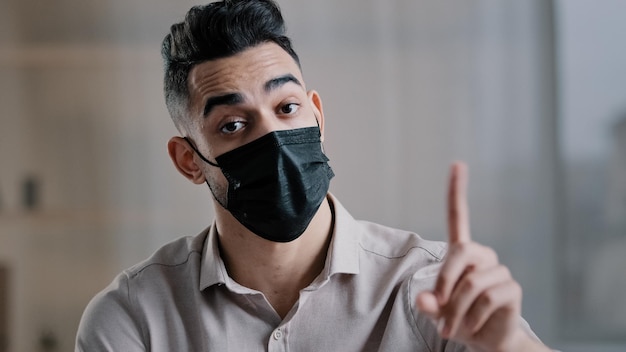 Empregado de empresário árabe usa máscara médica sentado na mesa de escritório balança o dedo indicador de onda alerta