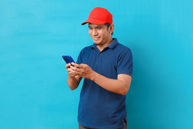 Foto empregado de entrega sorridente, homem asiático de boné vermelho, uniforme azul, roupas de trabalho, trabalho, conceito de serviço de revendedor de correio, segure na mão usando o celular