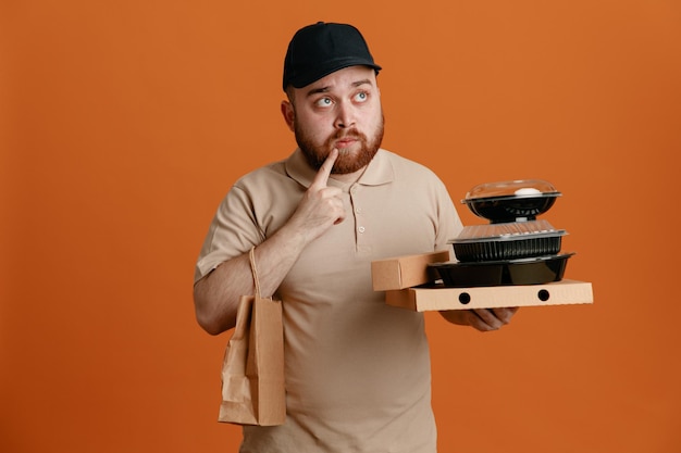 Empregado de entregador de boné preto e uniforme de camiseta em branco segurando recipientes de comida com saco de papel olhando para cima pensando confuso em pé sobre fundo laranja