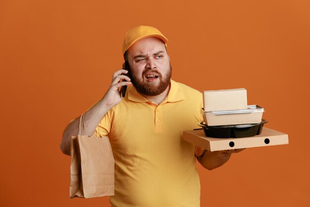 Empregado de entregador em uniforme de camiseta em branco de boné amarelo segurando o recipiente de comida e caixa de pizza com saco de papel falando no celular sendo descontente em pé sobre fundo laranja