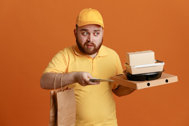 Empregado de entregador em uniforme de camiseta em branco de boné amarelo segurando recipiente de comida e caixa de pizza com saco de papel segurando dinheiro parecendo confuso em pé sobre fundo laranja