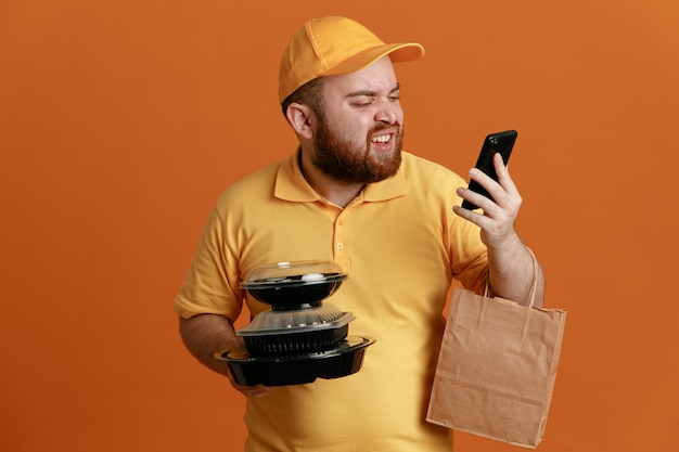 Empregado de entregador em uniforme de camiseta em branco de boné amarelo segurando recipientes de comida com saco de papel falando no celular com raiva e frustrado em pé sobre fundo laranja