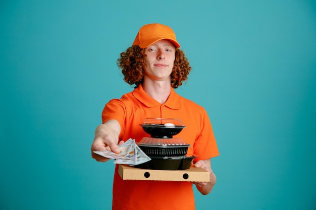 Empregado de entregador em uniforme de camiseta em branco de boné laranja segurando recipientes de comida olhando para câmera sorrindo amplamente feliz e positivo retornando a mudança em pé sobre fundo azul