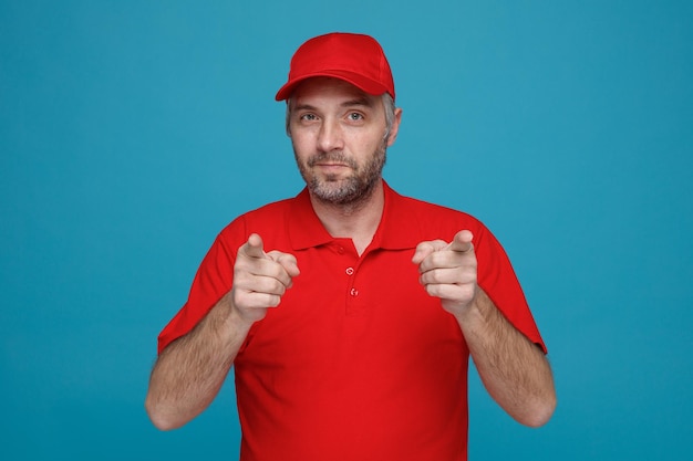 Empregado de entregador em uniforme de camiseta em branco de boné vermelho apontando com o dedo indicador para a câmera sorrindo confiante em pé sobre fundo azul