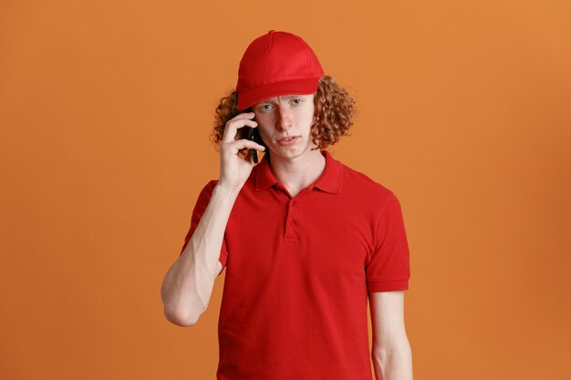 Empregado de entregador em uniforme de camiseta em branco de boné vermelho falando no celular olhando para a câmera com expressão triste em pé sobre fundo laranja