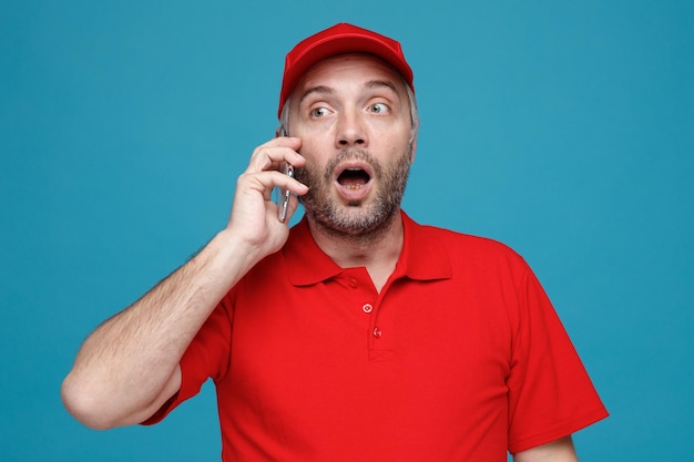 Empregado de entregador em uniforme de camiseta em branco de boné vermelho falando no celular parecendo espantado e surpreso em pé sobre fundo azul