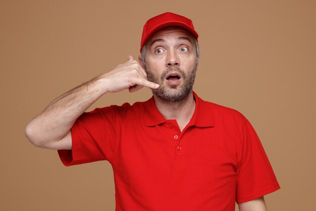 Empregado de entregador em uniforme de camiseta em branco de boné vermelho fazendo me chamar de gesto parecendo espantado e surpreso em pé sobre fundo marrom