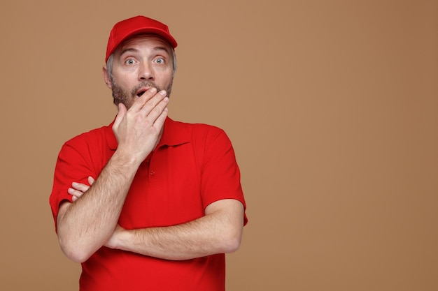 Empregado de entregador em uniforme de camiseta em branco de boné vermelho olhando para a câmera sendo chocado cobrindo a boca com a mão em pé sobre fundo marrom