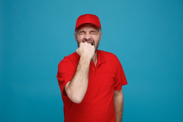 Empregado de entregador em uniforme de camiseta em branco de boné vermelho parecendo estressado e nervoso mordendo o punho em pé sobre fundo azul