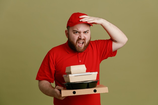 Empregado de entregador em uniforme de camiseta em branco de boné vermelho segurando recipientes de comida e caixa de pizza olhando para a câmera com cara feia gritando e gritando em pé sobre fundo verde