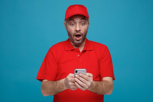 Empregado de entregador em uniforme de camiseta em branco de boné vermelho segurando smartphone olhando para a tela sendo surpreendido e espantado em pé sobre fundo azul