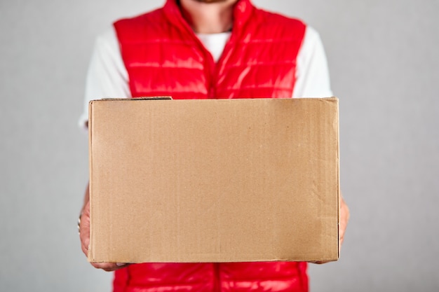 Empregado de entregador em uniforme de colete vermelho segura caixa de papelão vazia isolada na parede cinza, coronavírus pandêmico de quarentena de serviço, entrega sem contato.