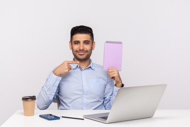 Empregado de escritório homem elegante sentado no local de trabalho, apontando a folha vazia e sorrindo para a câmera, mostrando o modelo de caderno de papel, simulando espaço em branco para ideia de negócio, mensagem. tiro de estúdio isolado