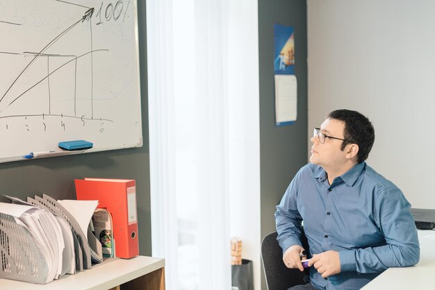 Empregado de homem de camisa azul e óculos sentado no escritório assistindo no gráfico no quadro branco