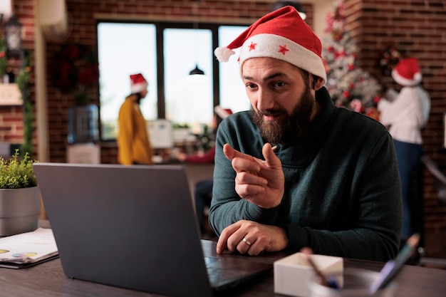 Empregado do sexo masculino falando em reunião de chamada de videoconferência no escritório de negócios com decorações festivas e árvore de natal. Usando teleconferência online remota para conversar durante a temporada de férias.