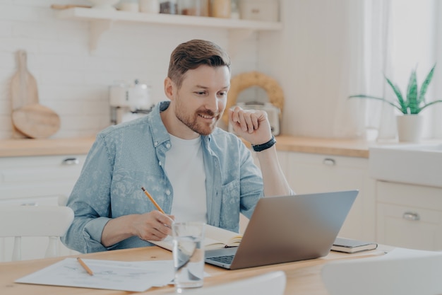 Empregado do sexo masculino sorridente usando o laptop em casa, sentado à mesa da cozinha, olhando para a tela e fazendo videochamada ou assistindo ao webinar, escrevendo notas no caderno. Estudo à distância e conceito freelance