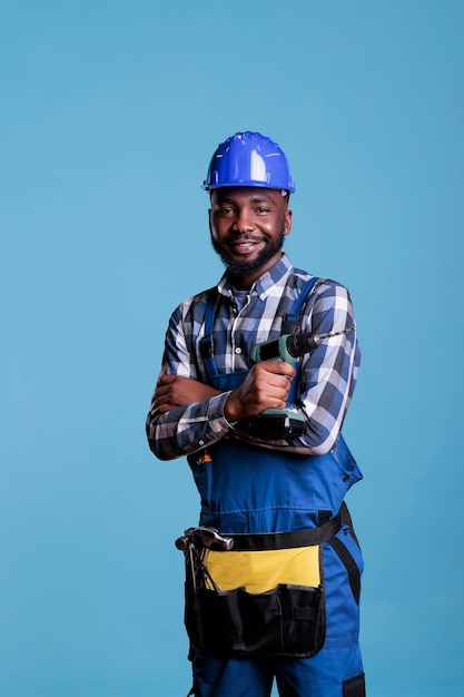 Empreiteiro profissional com capacete e cinto de ferramentas sobre fundo azul claro. Construtor afro-americano segurando furadeira sem fio usando faixa de cintura enquanto olha sorrindo para a câmera.