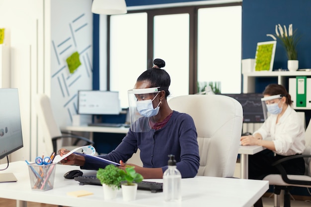 Emprendedor africano haciendo análisis financiero con mascarilla durante la pandemia de coronavirus. Compañeros de trabajo multiétnicos que trabajan respetando la distancia social en la empresa financiera.