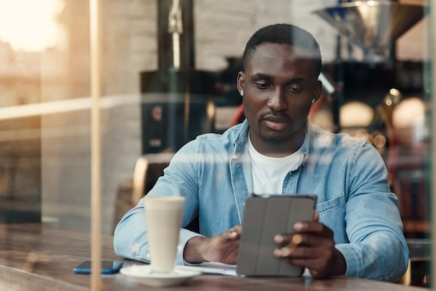 Un emprendedor negro con propósito usa tablet pc mientras está sentado en la cafetería con café cerca de la ventana