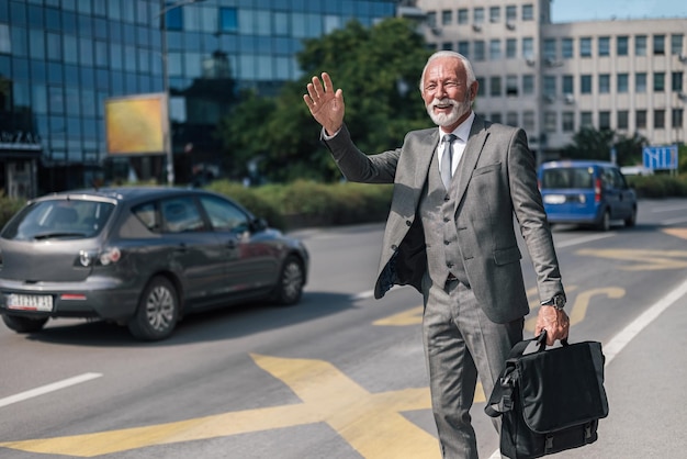 Emprendedor senior con bolsa de computadora portátil llamando a un taxi en la ciudad durante el día soleado