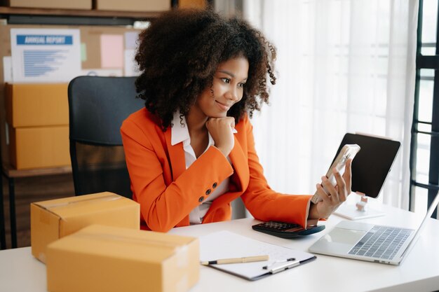 Foto emprendedora africana que utiliza un teléfono inteligente o una tableta para recibir y comprobar compras en línea, pedidos de compras, preparar cajas de productos en su oficina en casa.