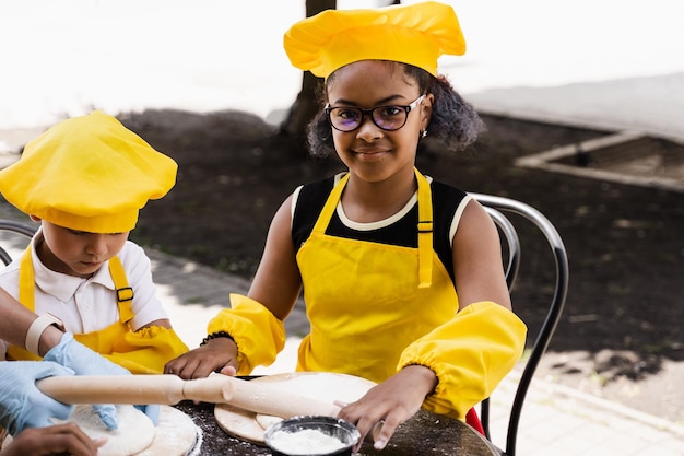 Empresa multinacional de niños cocineros con uniformes amarillos cocinando masa para panadería Adolescente africano y niña negra se divierten con un niño caucásico y cocinan comida