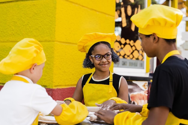 Empresa multinacional de niños cocineros con uniformes amarillos cocinando masa para panadería Adolescente africano y niña negra se divierten con un niño caucásico y cocinan comida