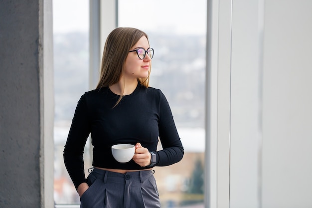 Empresaria adulta profesional tomando café en camisa y pantalón de pie delante de grandes ventanales.