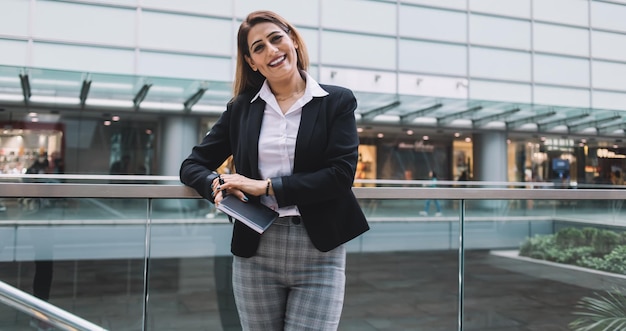 Empresária alegre segurando notebook e em pé perto de cerca de vidro perto de edifício moderno