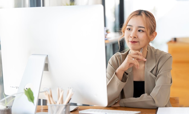 Empresária alegre trabalhando em um computador desktop no escritório Empresária asiática feliz e bonita em traje formal trabalhando no local de trabalho