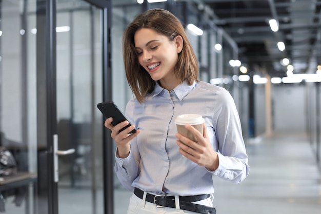 Empresária andando pelo corredor do escritório com copo de papel e smartphone.