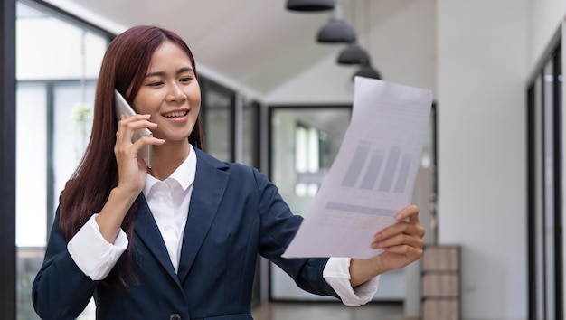 Empresária asiática atraente ou consultora financeira feminina lendo uma papelada de relatório financeiro e falando ao telefone com seu chefe