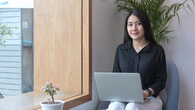 Empresária asiática atraente sentada na poltrona em seu café e usando o computador portátil