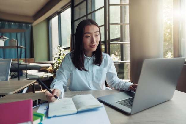 Empresária asiática atraente trabalhando no laptop e fazendo anotações no escritório moderno