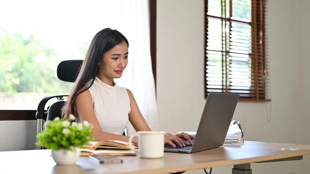 Empresária asiática bem-sucedida trabalhando em sua mesa em seu escritório moderno usando laptop