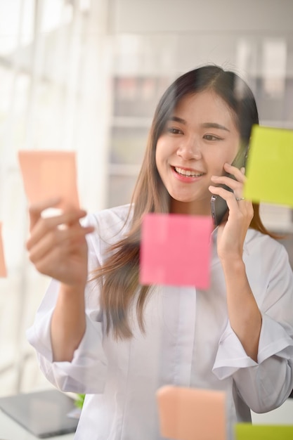 Empresaria asiática creativa leyendo detalles en notas adhesivas mientras habla por teléfono