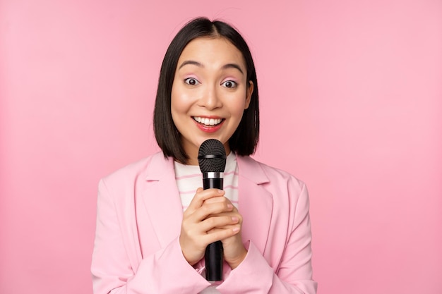 Empresária asiática dando discurso segurando o microfone e sorrindo de terno sobre fundo rosa