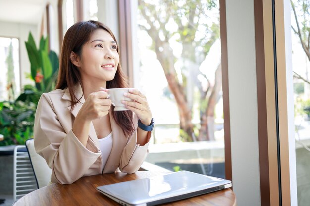 Empresária asiática de terno casual trabalhando e bebendo café por laptop na cafeteria