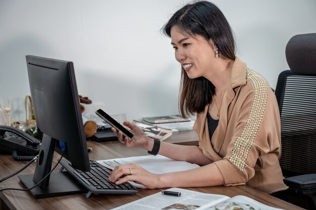 Empresária asiática em terno formal, escrevendo e trabalhando com computador e dispositivos móveis no escritório, negócios e plano de projeto para o conceito de serviço ao cliente