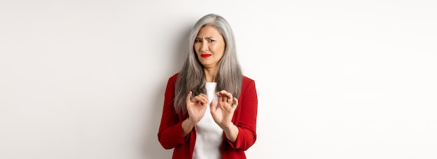 Empresária asiática enojada com cabelos grisalhos vestindo blazer vermelho e maquiagem rejeitando algo nojento