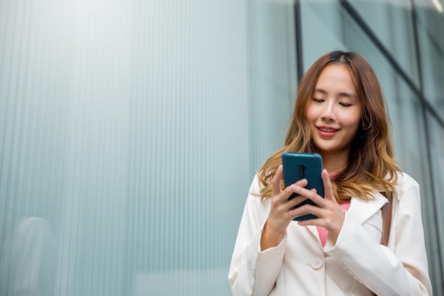 Empresária asiática enviando mensagens de texto para smartphone no trabalho, ela andando perto de seu prédio de escritórios pela manhã, Linda mulher de negócios sorrindo segurando o celular ao ar livre caminhando na rua da cidade urbana